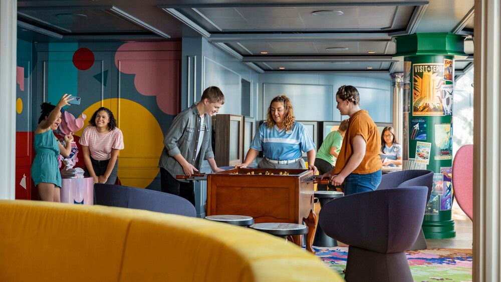 Three teenagers playing foosball inside Vibe, a teen club on board the Disney Adventure