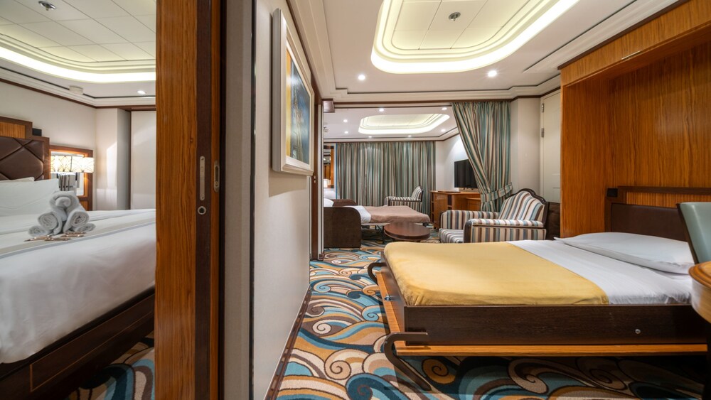 A bedroom with a bed, a nightstand, a sofa, 2 chairs and a TV in a Disney Dream Concierge 1 Bedroom Suite with Verandah