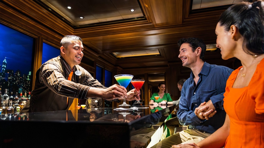 Um cast member servindo coquetéis para dois hóspedes no bar Skyline