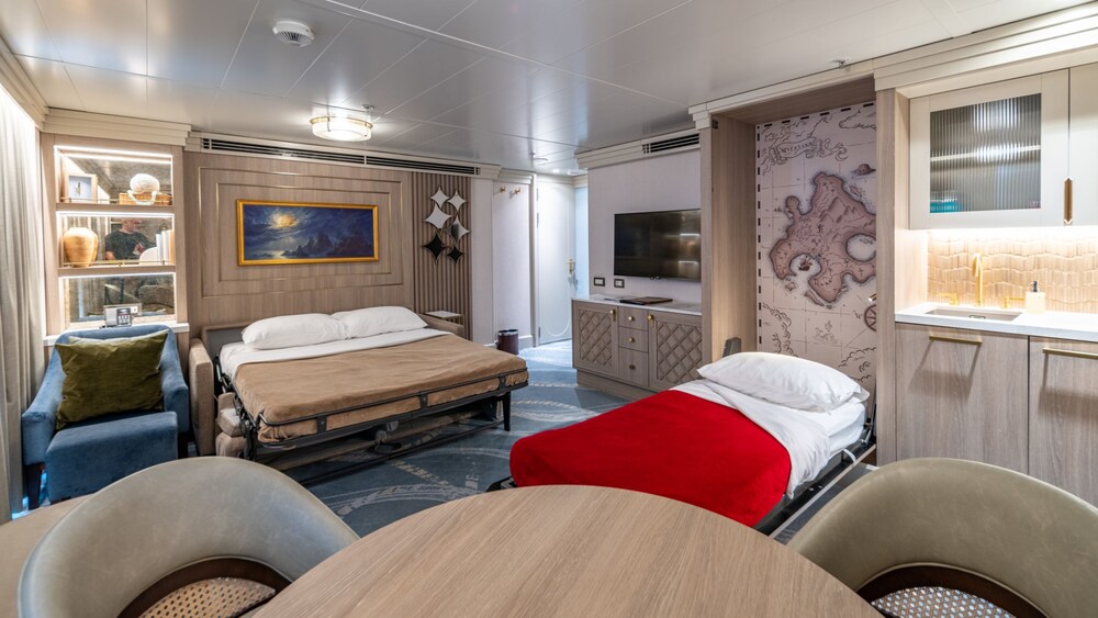 A living area with a wall bed, sofa bed, dining table, TV, wet bar and 3 chairs in a Disney Magic Concierge 1 Bedroom Suite with Verandah