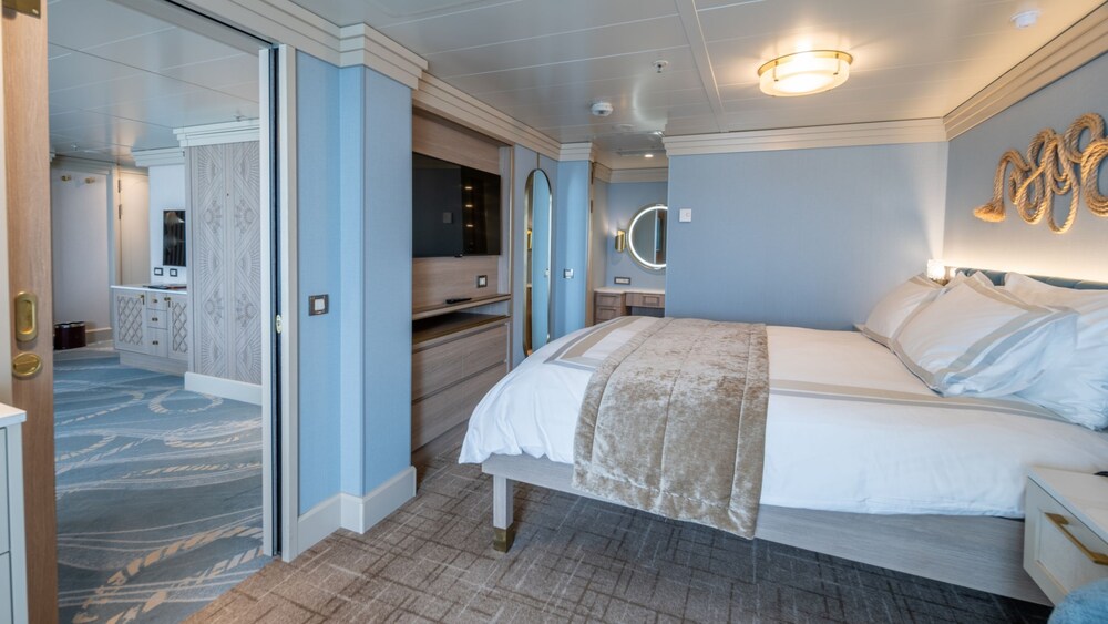 A bedroom with a bed, nightstand, TV, dresser and mirror in a Disney Magic Concierge 1 Bedroom Suite with Verandah