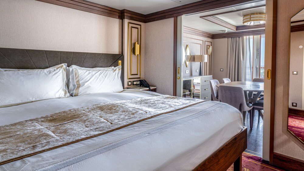 A bedroom with a bed, nightstand, mirror and an adjacent dining area in a Disney Magic Concierge Royal Suite with Verandah