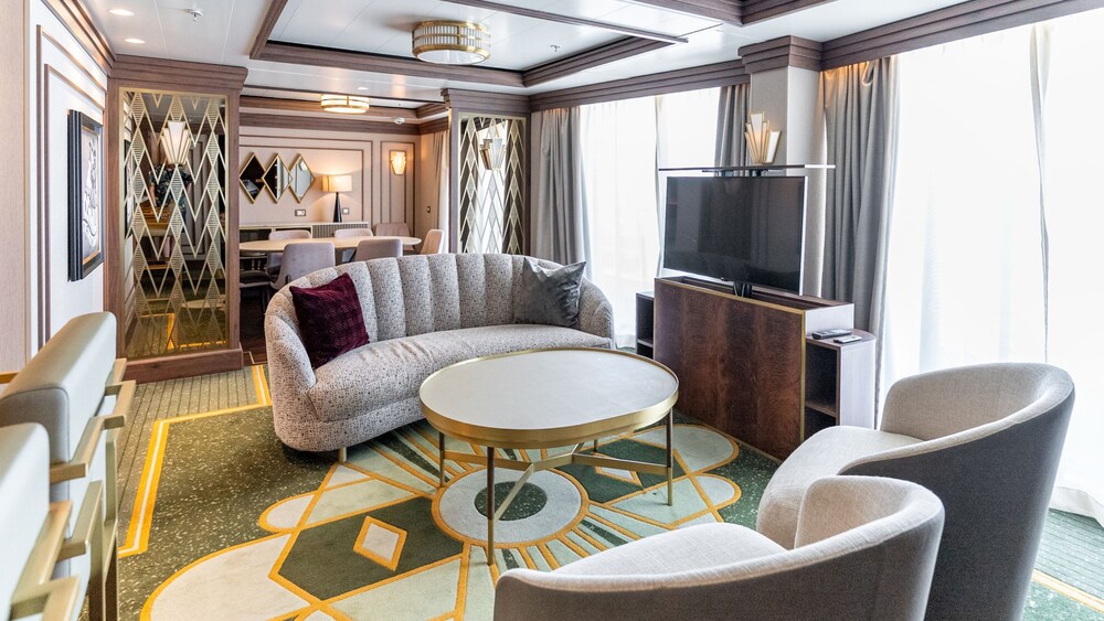 A living area with a sofa, table, TV, 2 chairs and an adjacent dining area in a Disney Magic Concierge Royal Suite with Verandah