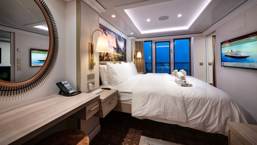 An artist rendering of a Disney Treasure Concierge 1 Bedroom Suite with Verandah Stateroom with a vanity, artwork inspired by The Lion King, a bed, a TV and a verandah with an ocean view 