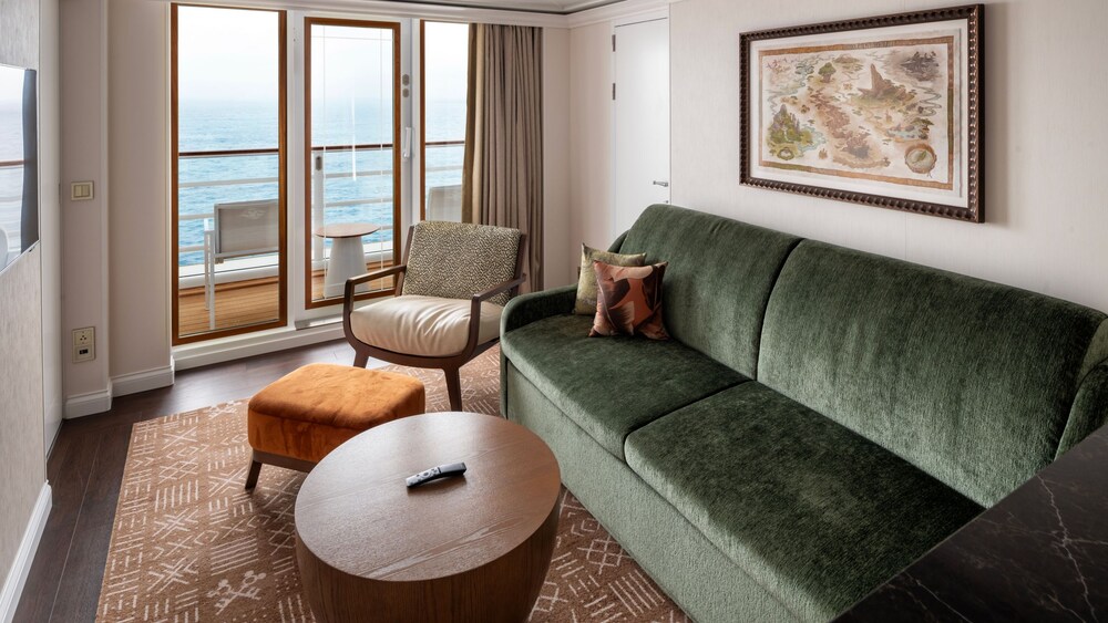 A living area with a sofa, chair, coffee table, TV, The Lion King artwork and a door to a private deck in a Concierge stateroom on the Disney Treasure
