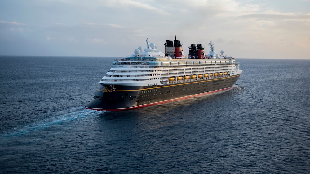 A Disney Cruise Line ship sailing across the open ocean, viewed from the front and starboard side