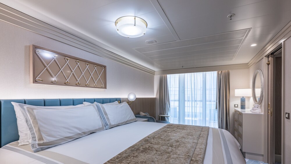 A bedroom with a bed, chair, dresser, mirror and verandah in a Disney Wonder Concierge 1 Bedroom Suite with Verandah