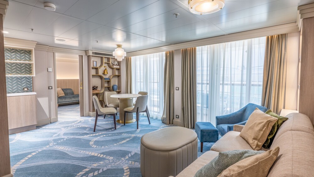 A living area with a sofa, chair, dining table with chairs and 2 ottomans in a Disney Wonder Concierge 1 Bedroom Suite with Verandah