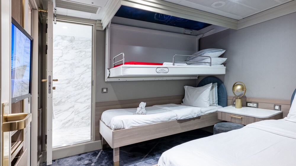 A bedroom with a TV, desk, stool, 2 beds and an adjacent bathroom in a Disney Wonder Concierge 2 Bedroom Suite with Verandah