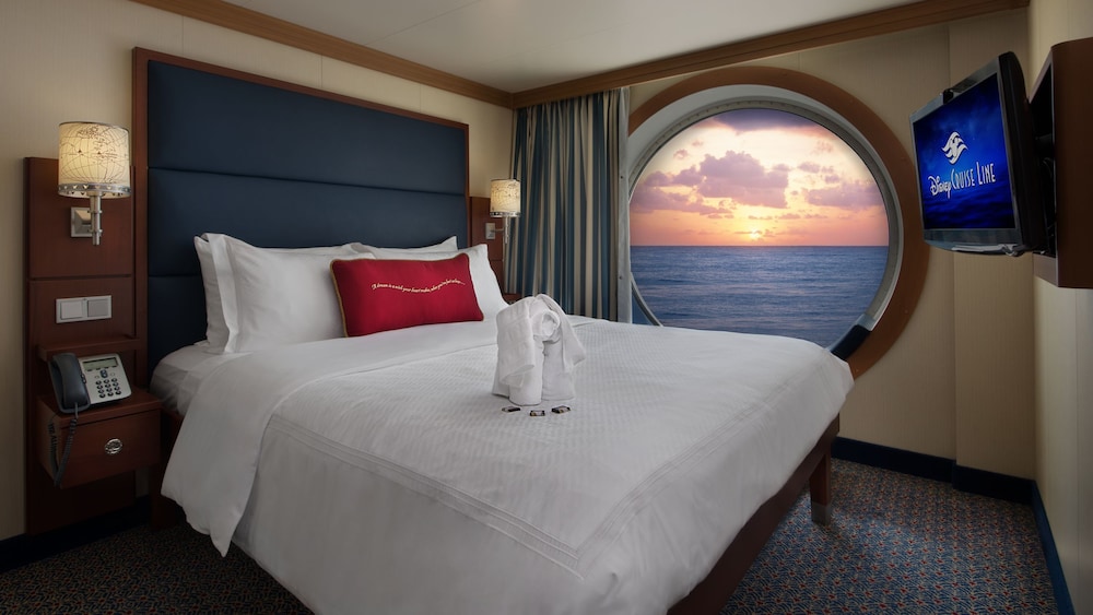 A Disney Cruise Line Deluxe Family Oceanview Stateroom with a queen bed, a sofa, a porthole with an ocean view, storage shelves, a desk and a TV