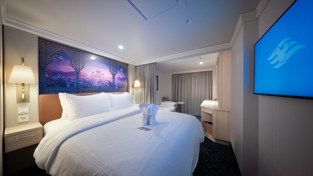 An artist rendering of a Disney Treasure Deluxe Oceanview with Verandah Stateroom with a bed, artwork inspired by Aladdin, a sofa, a verandah with an ocean view, a vanity and a TV