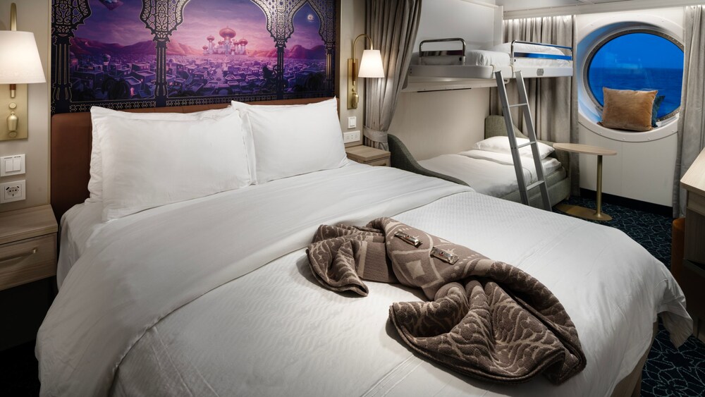A Disney Cruise Line Oceanview Stateroom with a folded blanket, bed, table, artwork inspired by Aladdin, 2 nightstands, 2 bunk beds and a window facing the ocean