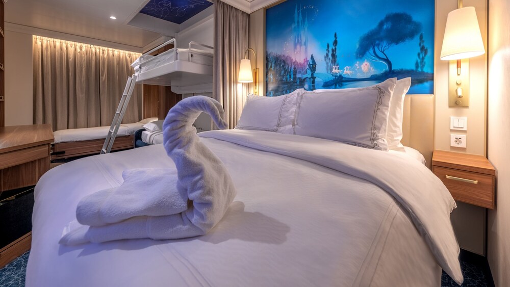A Disney Wish Deluxe Oceanview Stateroom with Verandah featuring a desk, artwork inspired by Cinderella, 2 nightstands, 2 beds, 2 bunk beds and a towel folded into the shape of a swan