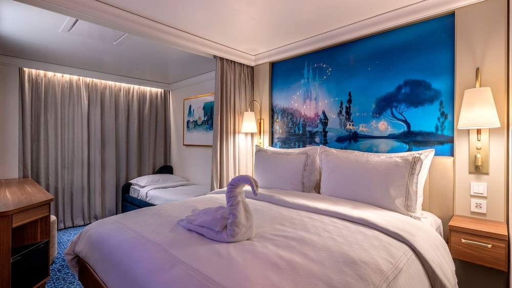 A Disney Wish Deluxe Oceanview Stateroom with a bed, nightstand, sofa bed, desk, artwork inspired by Cinderella and a towel folded into the shape of a swan