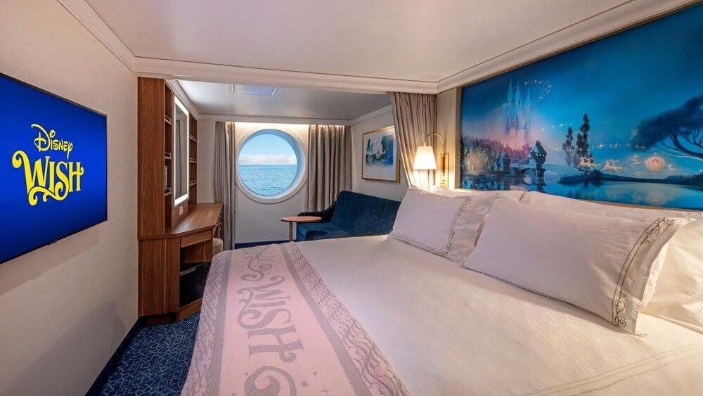 Un Camarote de Lujo con Vista al Mar del Disney Wish, con cama, sofá, mesa, escritorio, espejo, televisión, obras de arte inspiradas en Cenicienta y una ventana con vista al mar