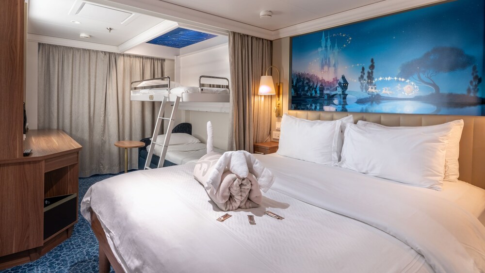 A Disney Wish Deluxe Oceanview Stateroom with a bed, sofa, table, desk, mirror, TV, artwork inspired by Cinderella and a window facing the ocean