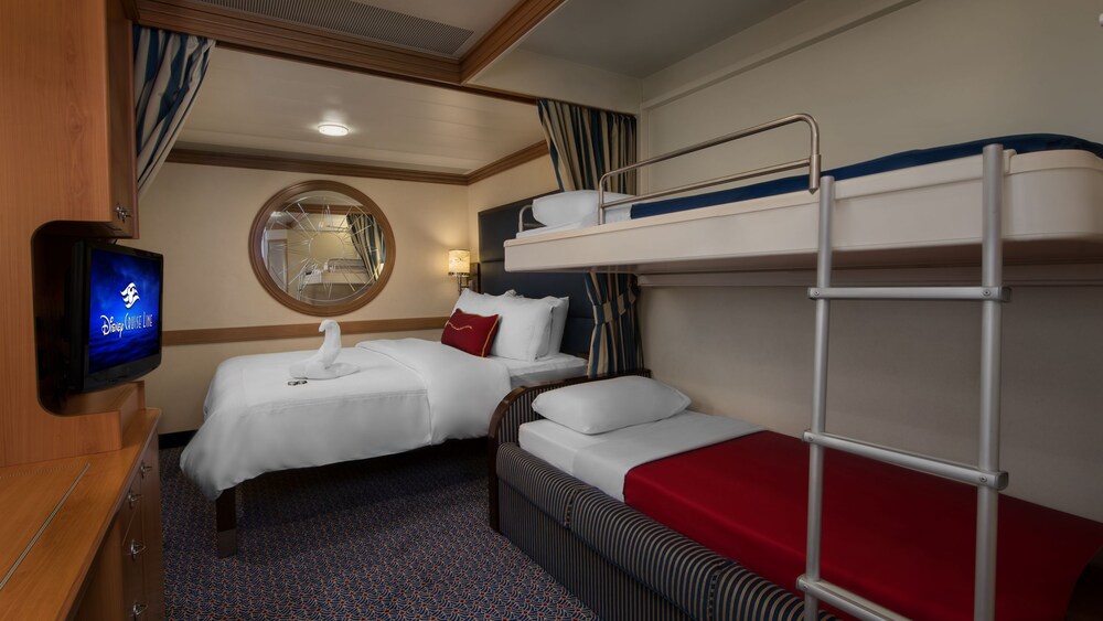 A Disney Cruise Line Deluxe Inside Stateroom with a bed, mirror, TV, desk, 2 bunk beds and a towel folded into the shape of a swan