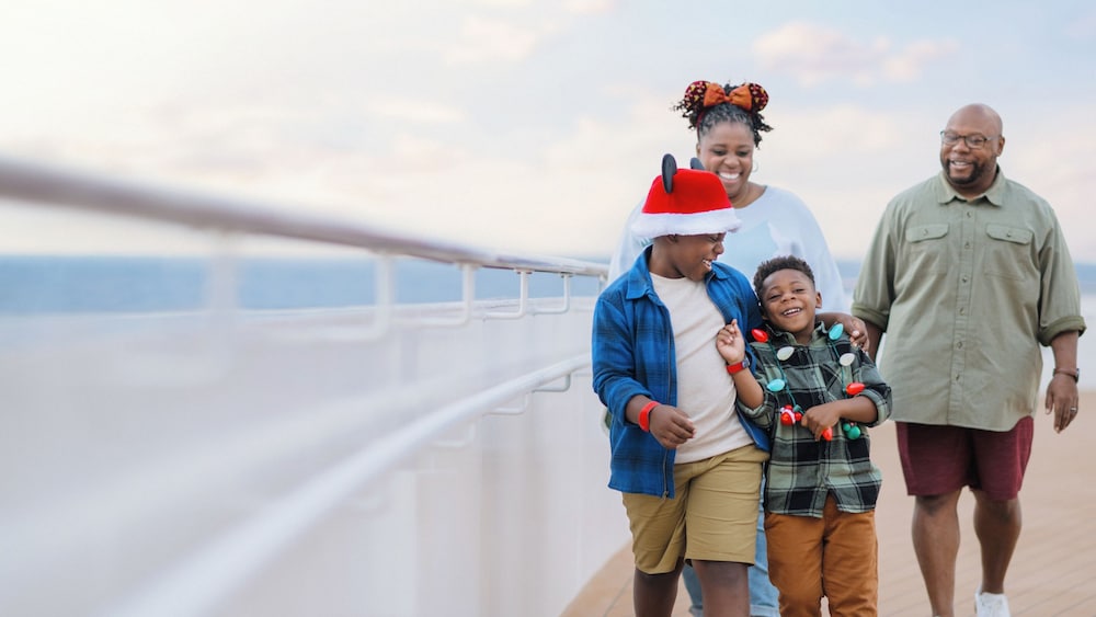 A family of 4, dressed in Christmas attire, smiling and interacting along the deck of a Disney Cruise Line ship