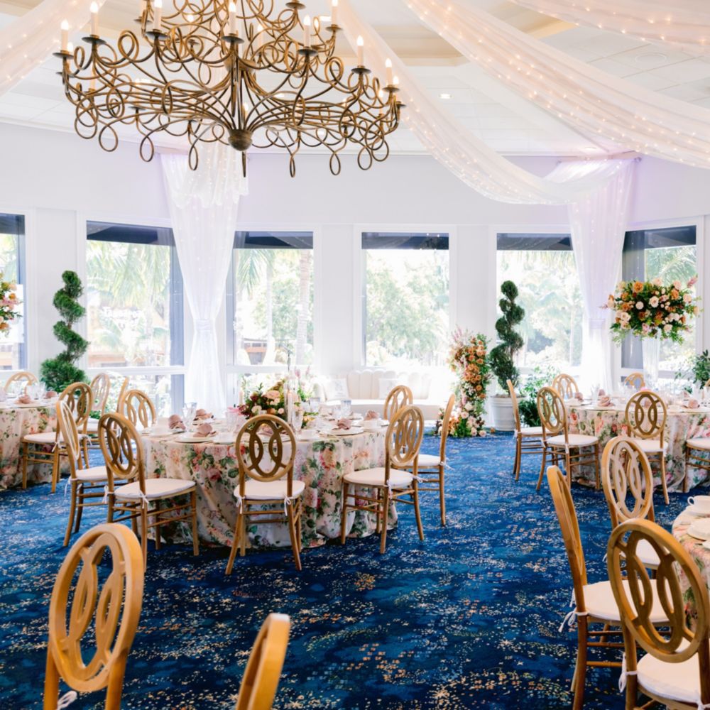 Un salón de baile con una lámpara de techo, mesas de comedor, arreglos florales, podas ornamentales, cubiertos y grandes ventanas con vistas al jardín