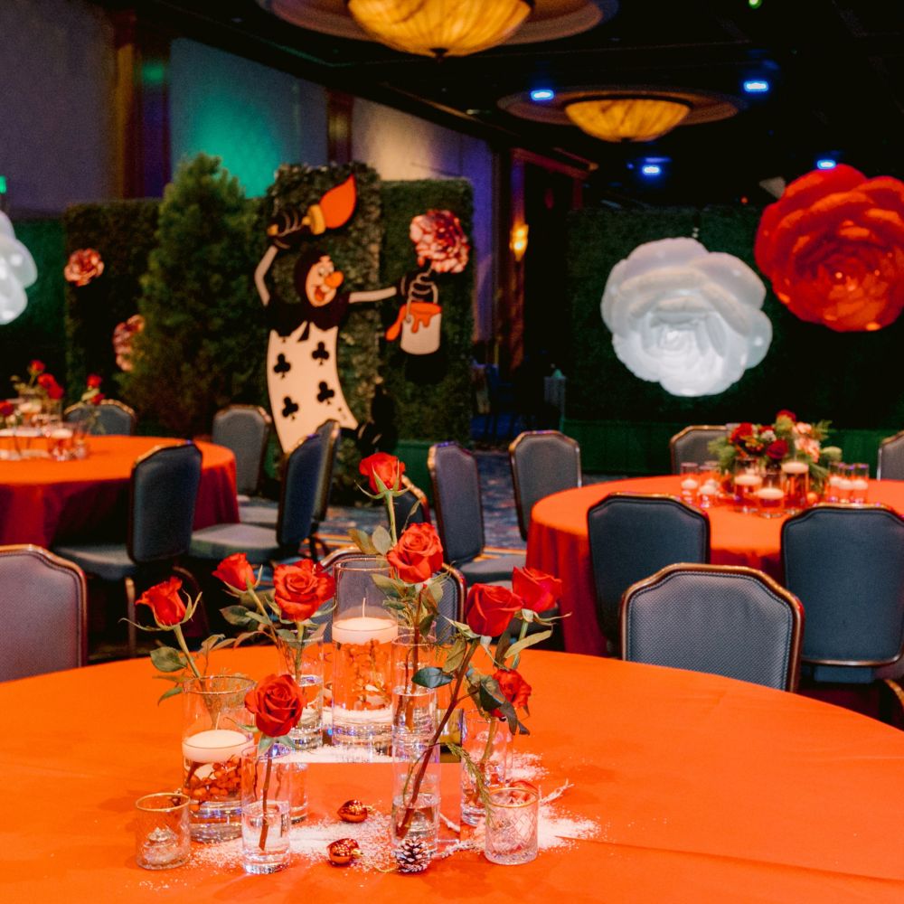 Un espacio para eventos con salón de baile con mesas, sillas, arreglos florales y decoraciones que recrean el jardín de rosas de Queen of Hearts de Alice in Wonderland