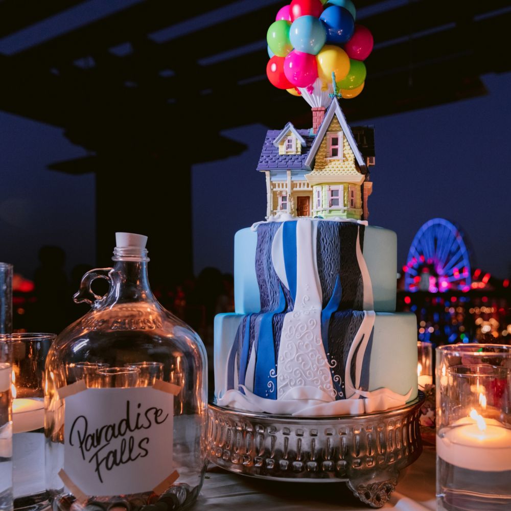 Una mesa con velas flotantes, una jarra de vidrio con una etiqueta que dice “Paradise Falls” y un pastel decorado con una cascada y la casa voladora de la película “Up” 