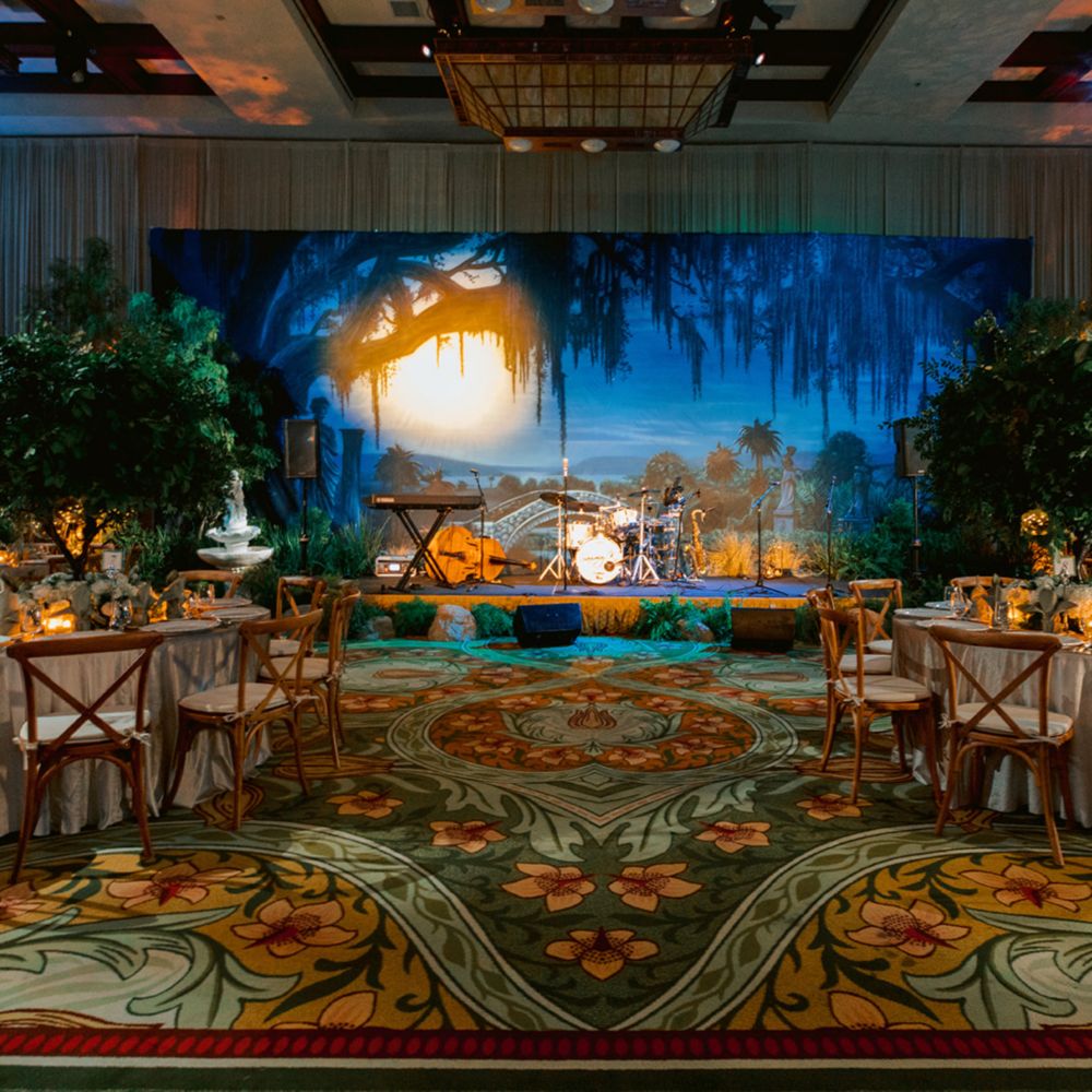 Un lugar para eventos con salón de baile con mesas de comedor, sillas, arreglos florales y un escenario con instrumentos musicales y un fondo de pantanos flanqueado por árboles