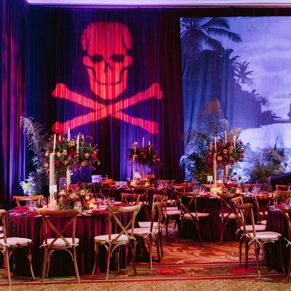 Un lugar para eventos con salón de baile con mesas de comedor, sillas, velas, arreglos florales y un telón de fondo con un barco pirata en una bahía y una proyección de una calavera y huesos cruzados