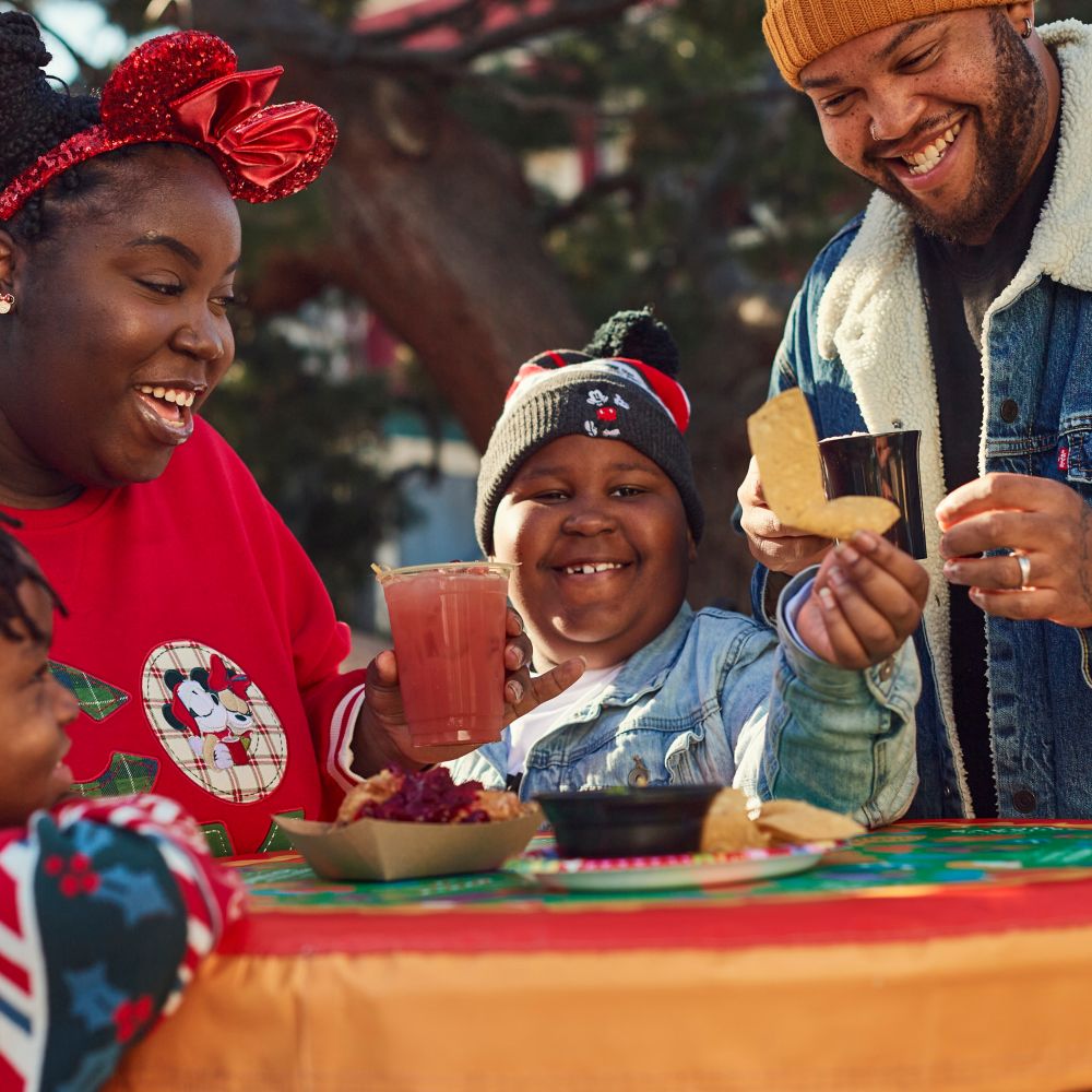Una familia con trajes navideños sentada en una mesa disfrutando de comidas y bebidas de la temporada de fiestas