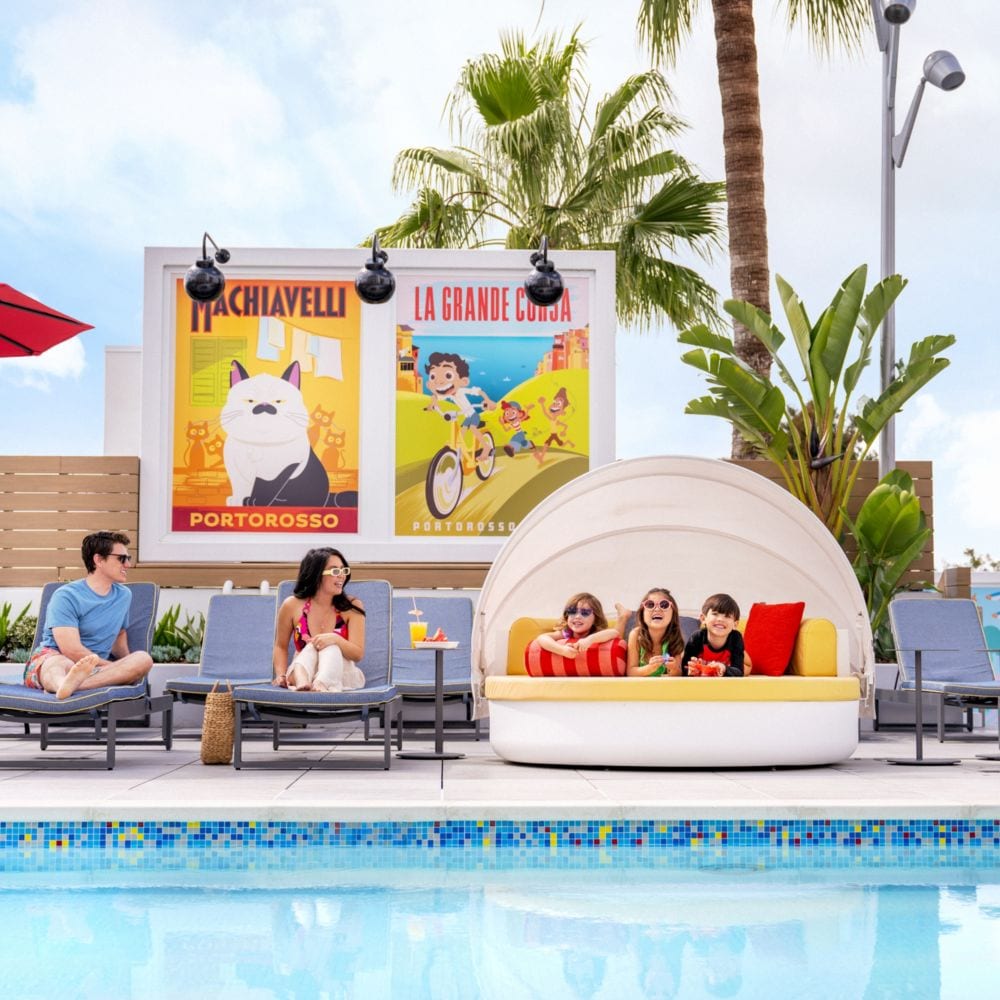 En el área de la piscina Pixel Pool de la azotea de Pixar Place Hotel, adultos sentados en camastros y tres niños relajándose en una cama de día, rodeados de llamativas obras de arte que representan historias de Pixar