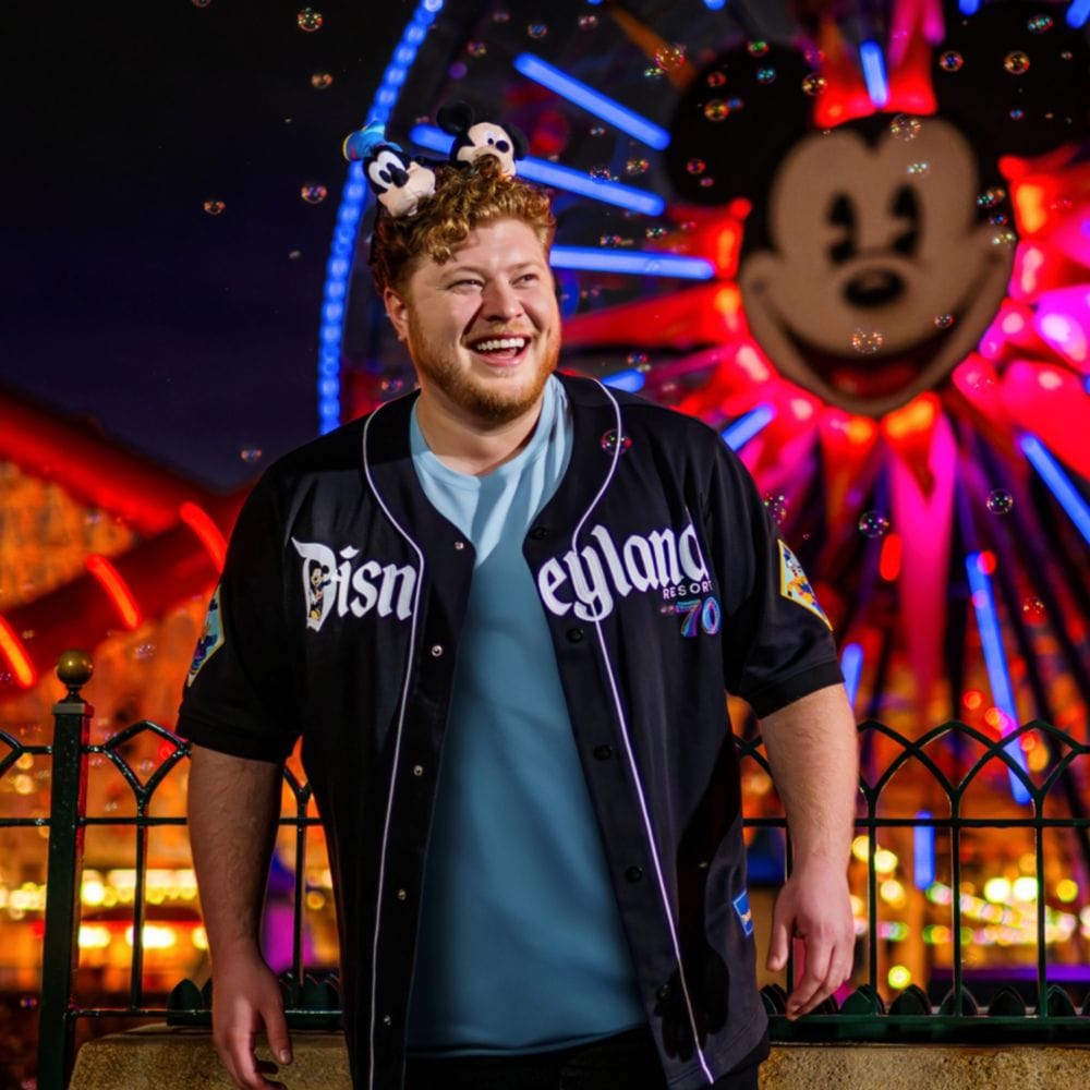 Visitante con una camiseta de béisbol del 70.º Aniversario de Disneyland y una diadema con orejas de Mickey Mouse, de pie junto a una barandilla con una noria iluminada que tiene el rostro de Mickey detrás, por la noche