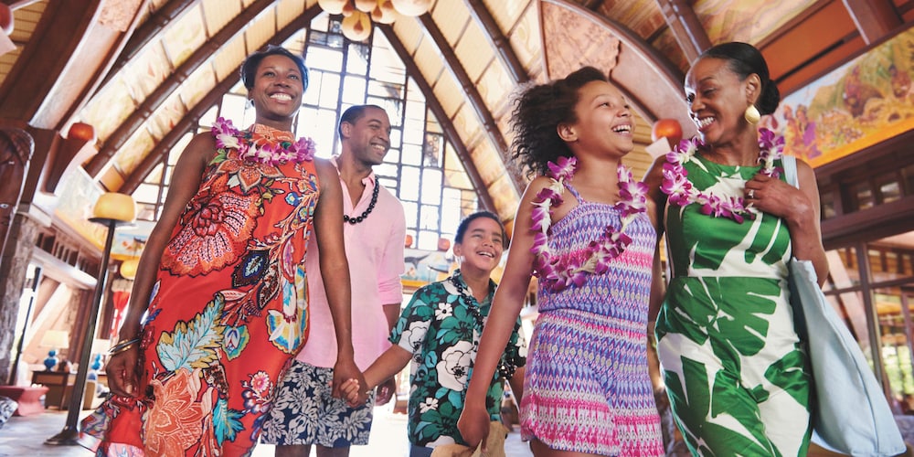 A multi-generational family smile and laugh as they walk through the main lobby at Aulani Resort wearing flower and kukui nut leis
