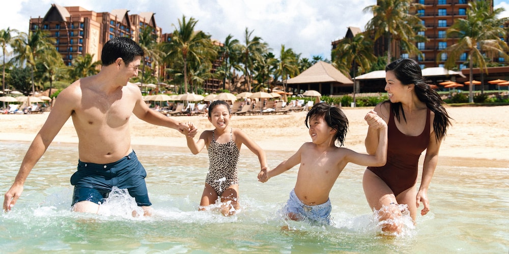 A father, mother, son and daughter holding hands as they wade in the lagoon at Aulani, A Disney Resort & Spa