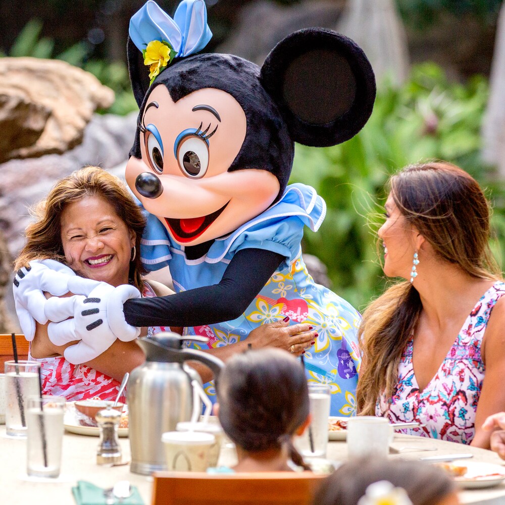 キャラクター・ダイニングで、耳に花を飾り笑顔の女性の膝に座るミニーマウス