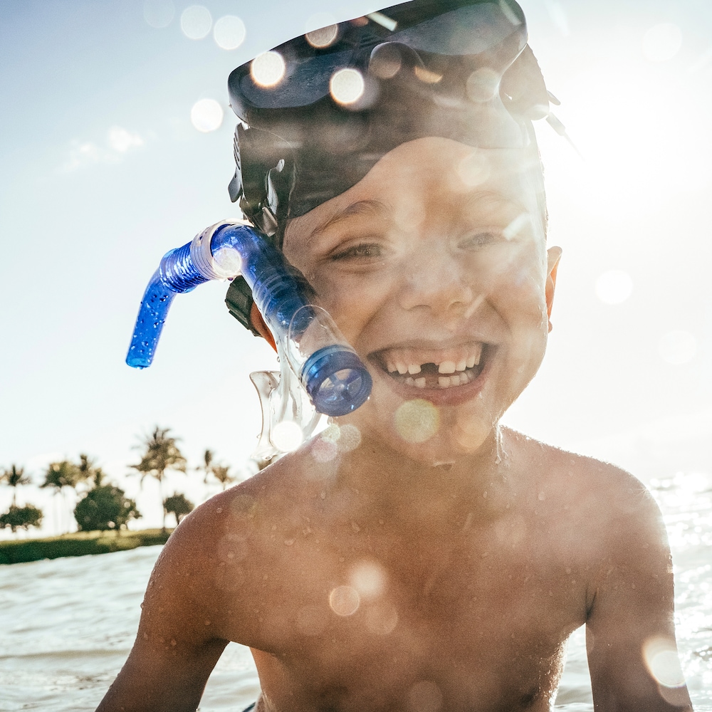 A smiling young boy in shallow ocean water, his snorkeling mask worn high on his forehead