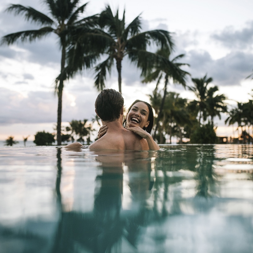 Two smiling adults in an infinity pool, with palms silhouetted along the shoreline