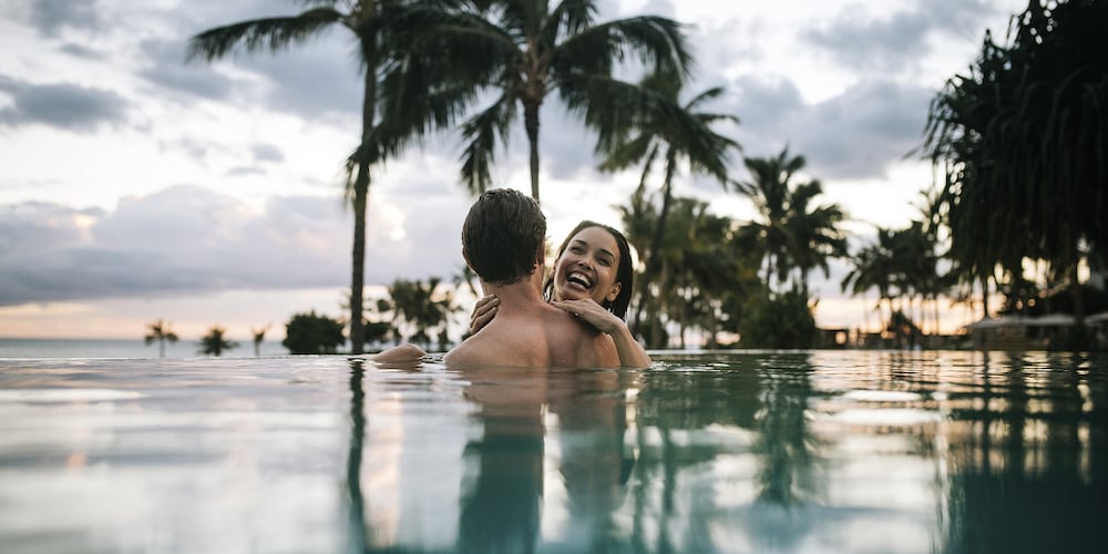 Two smiling adults in an infinity pool, with palms silhouetted along the shoreline
