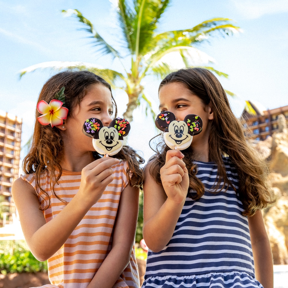 Two little girls look at each other as they eat their Mickey Mouse lollipops