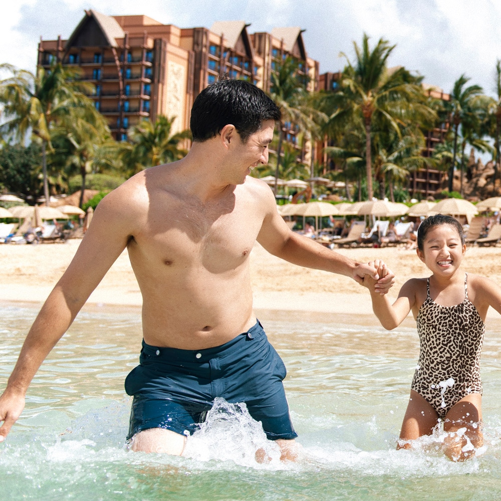 A father, mother, son and daughter holding hands as they wade in the lagoon at Aulani, A Disney Resort & Spa