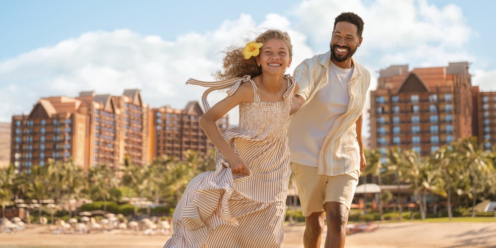 A girl and her father smiling as they run on the beach at Aulani Resort