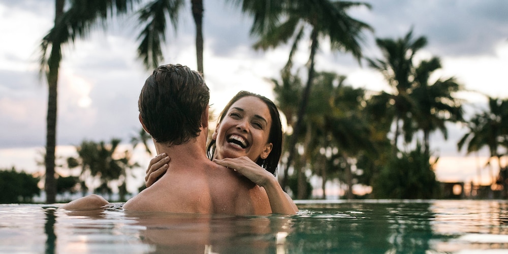 Two smiling adults in an infinity pool, with palms silhouetted along the shoreline