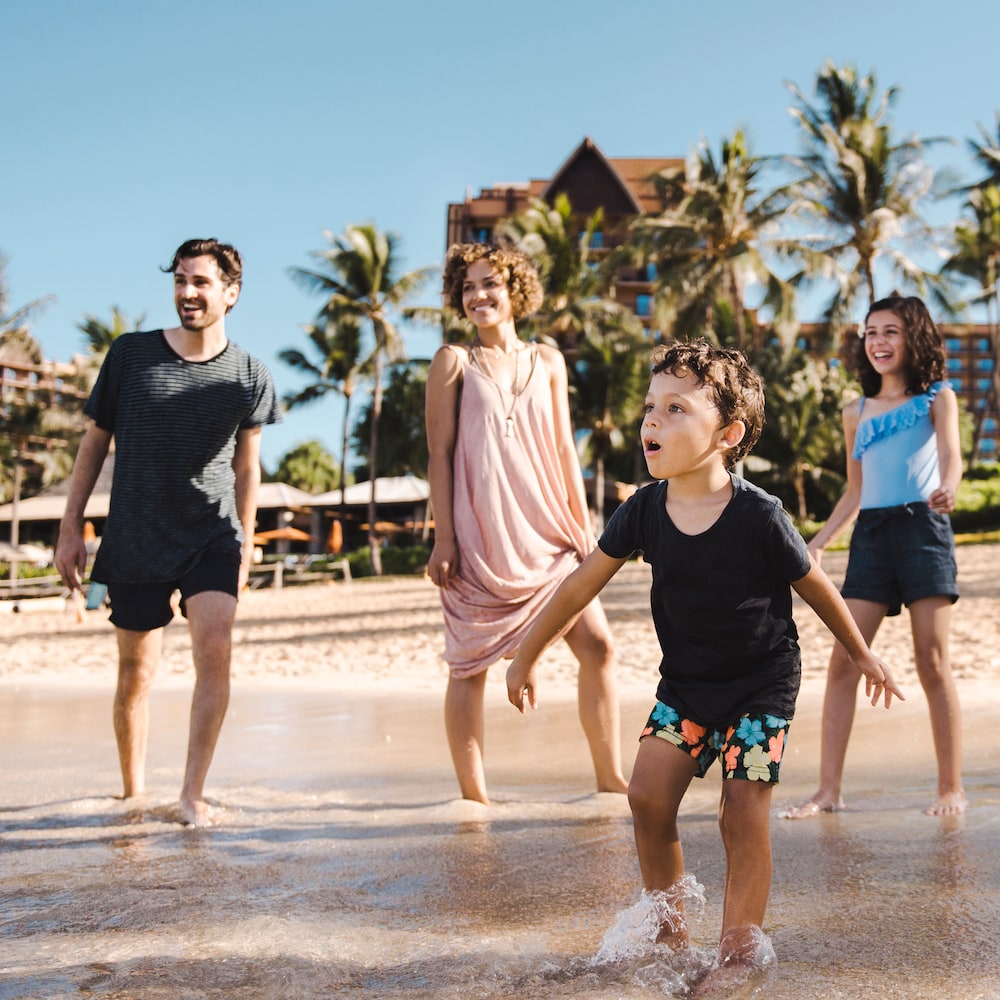 A family of 4 looks toward the ocean as they walk in the shallow water on the beach at Aulani Resort