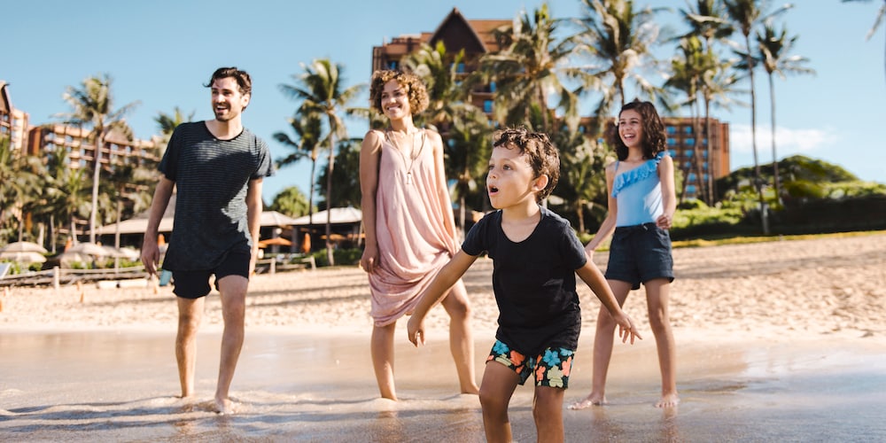 A family of 4 looks toward the ocean as they walk in the shallow water on the beach at Aulani Resort