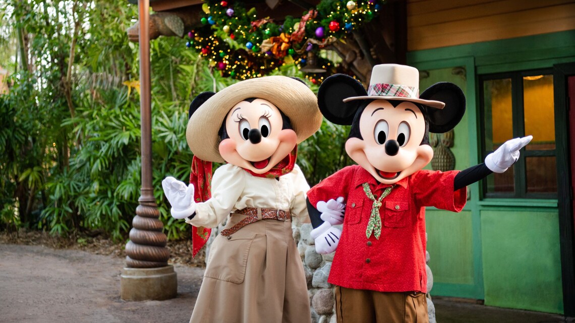 Mickey & Minnie wear Christmas themed adventure outfits standing in front of a wreath decoration at Adventurers Outpost