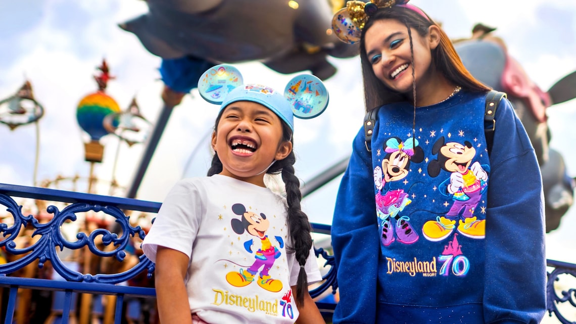 Dos Visitantes con vestimenta de la Celebración del 70.º Aniversario del Disneyland Resort, que incluye un sombrero de orejas de Mickey, una camiseta y una sudadera, parados frente a la atracción Dumbo the Flying Elephant