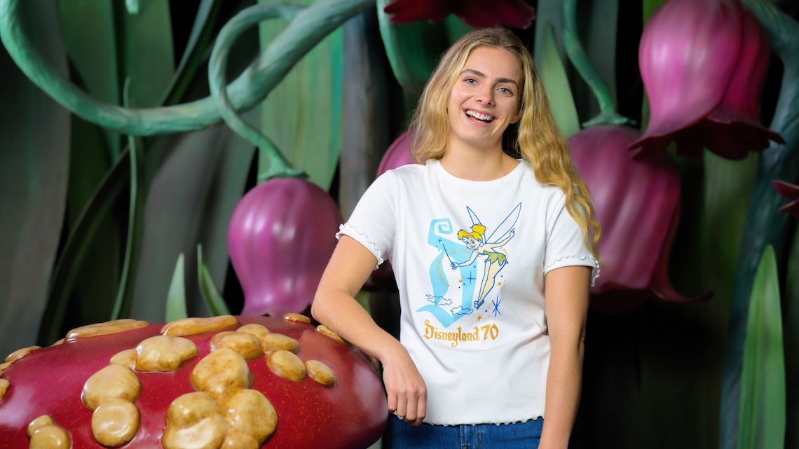 Una mujer sonriente con una camisa de la colección Tinker Bell del 70.º Aniversario de Disneyland mientras se apoya sobre un hongo gigante