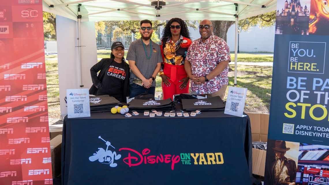 Four recruiters stand behind a Disney on the Yard information booth with promotional materials at an outdoor career event