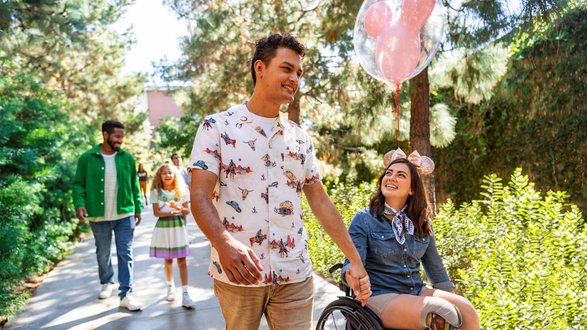 A man and a woman in a wheelchair on a tree lined path with a Mickey Mouse balloon