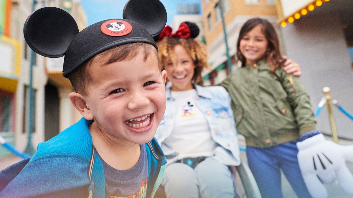 Dos niños y un Visitante en silla de ruedas sonriendo con un sombrero con orejas de Mickey Mouse, una diadema con orejas de Minnie Mouse y guantes de Mickey Mouse