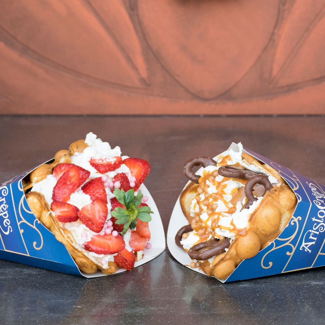 Two bubble waffles in Aristocrepes wrappers laying side by side, one is filled with strawberries and whipped cream, the other with chocolate ice cream, chocolate covered pretzels and whipped cream swirled with caramel