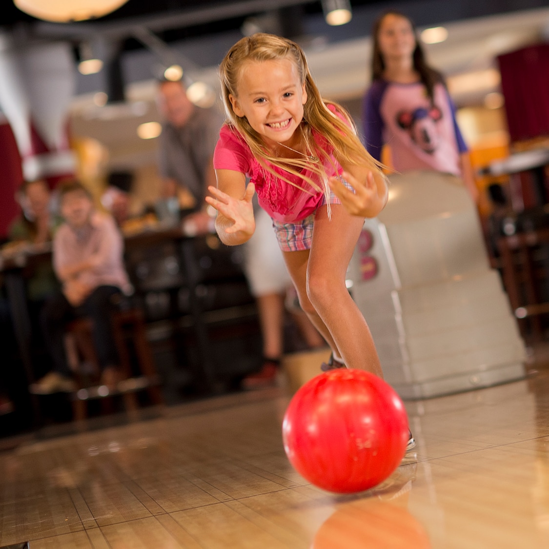 A smiling young girl bowls with both hands while her family watches in the background at Splitsville Luxury Lanes in Downtown Disney Area